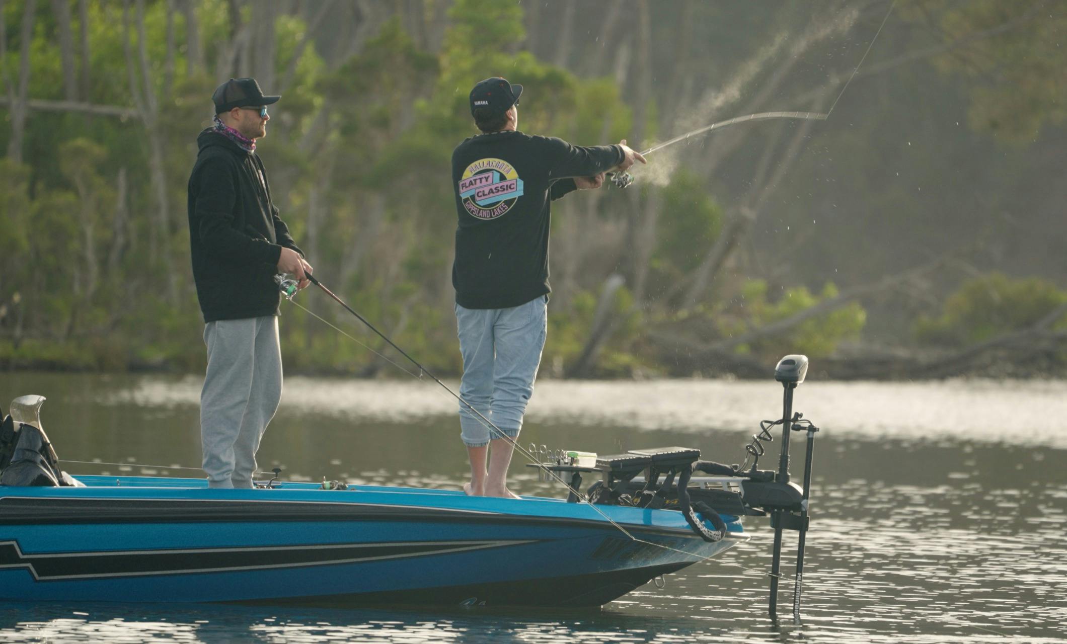 Angler in a Flatty Classic jumper casting from the bow of a blue boat near a timbered shoreline