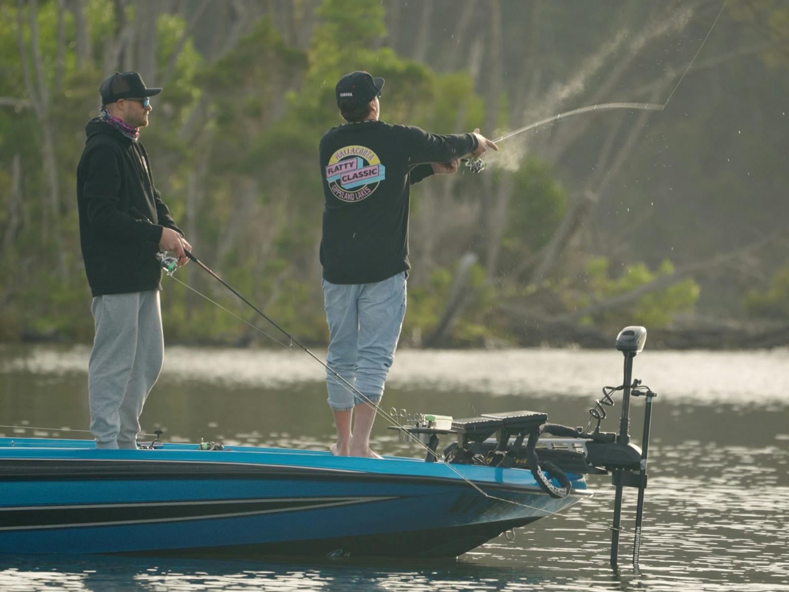 Angler in a Flatty Classic jumper casting from the bow of a blue boat near a timbered shoreline