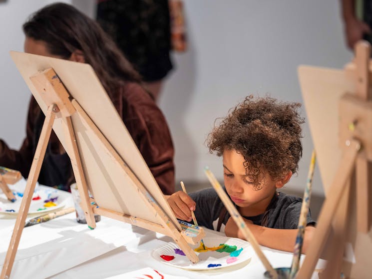 A young boy with curly hair paints a canvas on a small easel