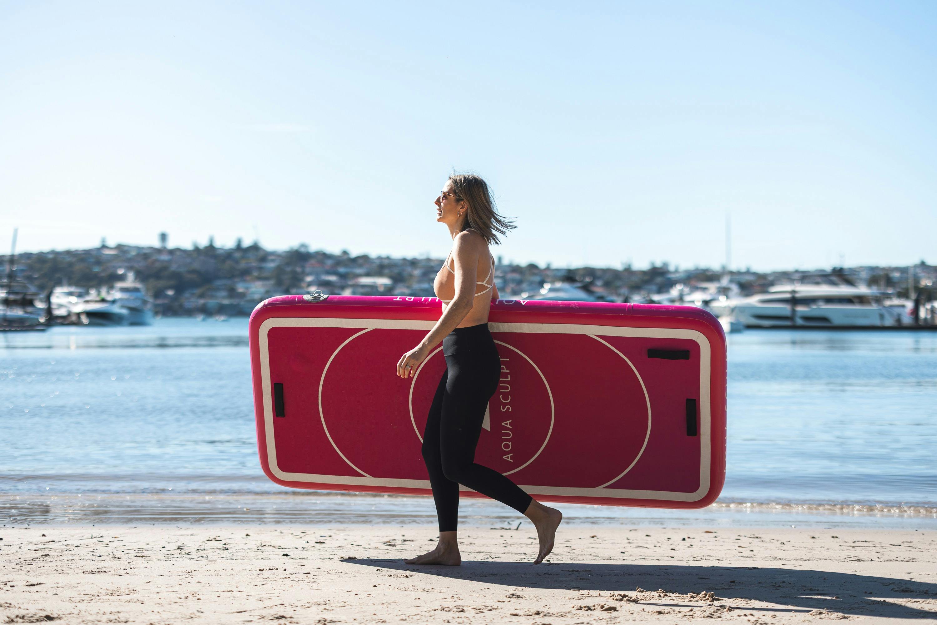 창립자 니나 허쉬펠트가 시드니 로즈 베이의 벨라미 해변을 따라 걷고 있으며 아쿠아 스컬프트 보드를 들고 있습니다.