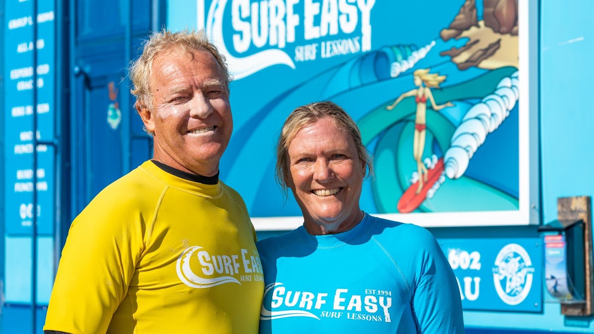 Man and woman smiling in front of surf school van.