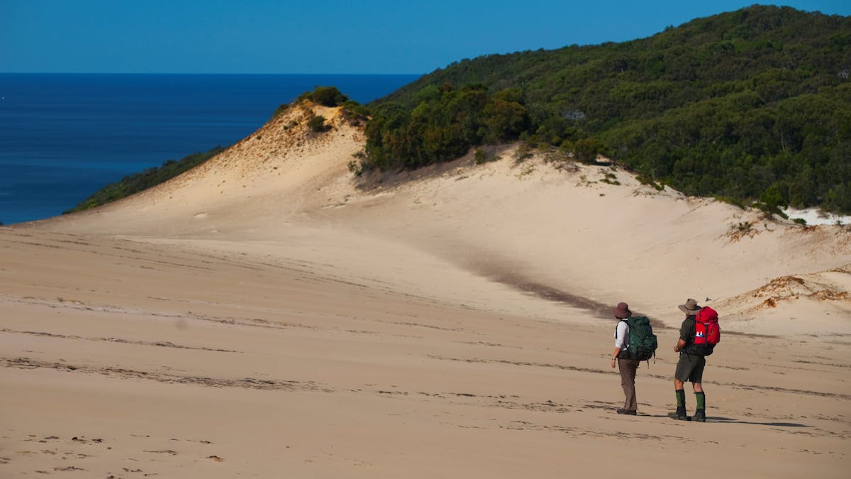 Two people with backpacks walking across a sand patch with ocean in the background.