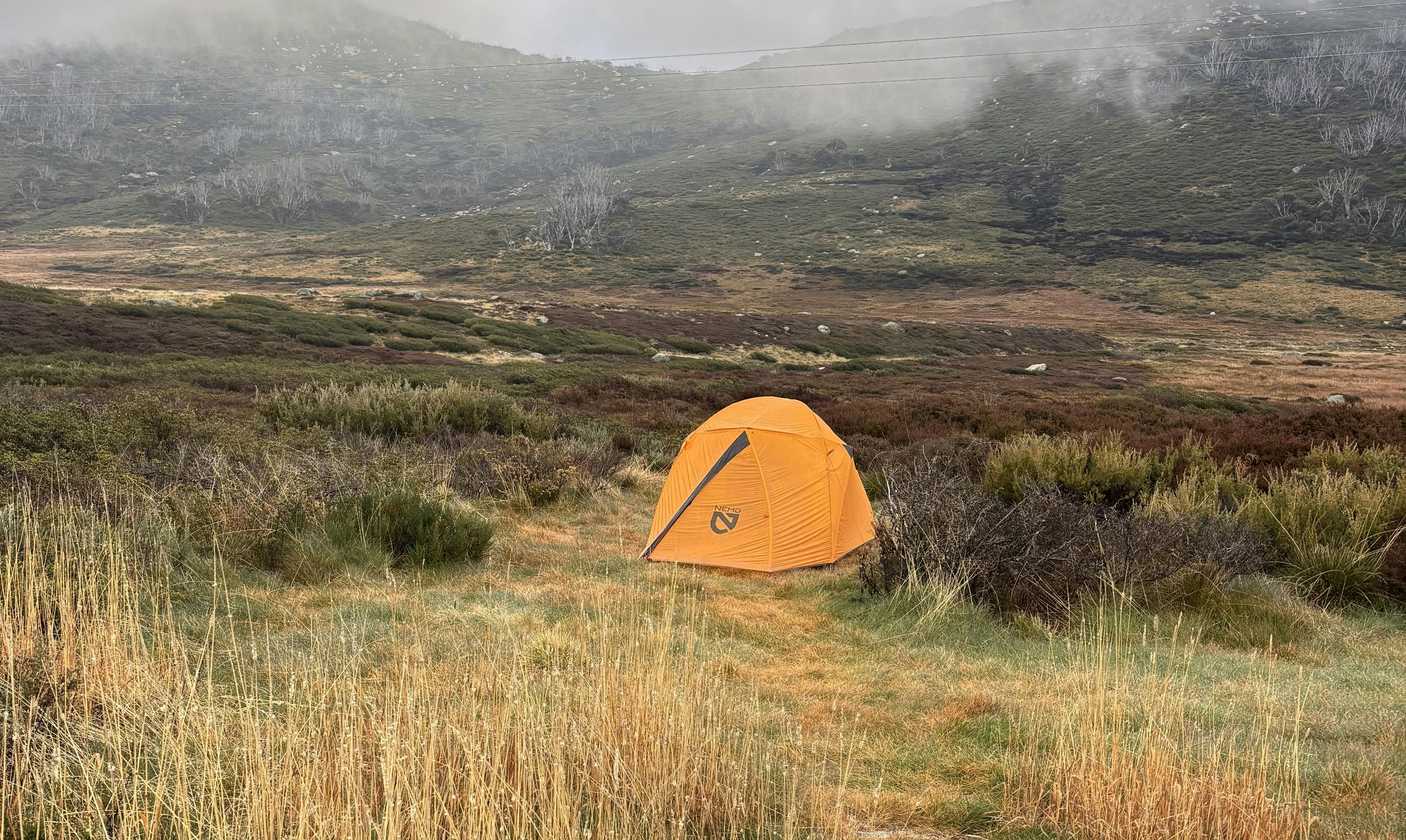 A small orange tent in the foreground against a background of rocky mountains and snow gums.