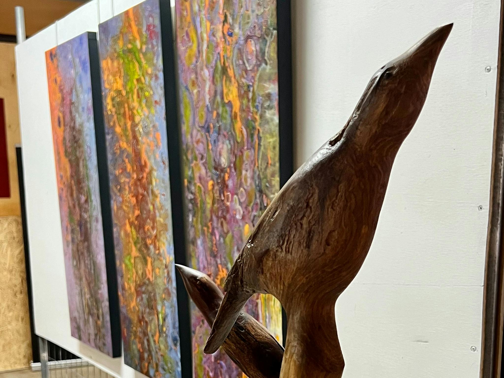 Hand carved wooden birds i on stand in front of a nanging triptych of bark paintings