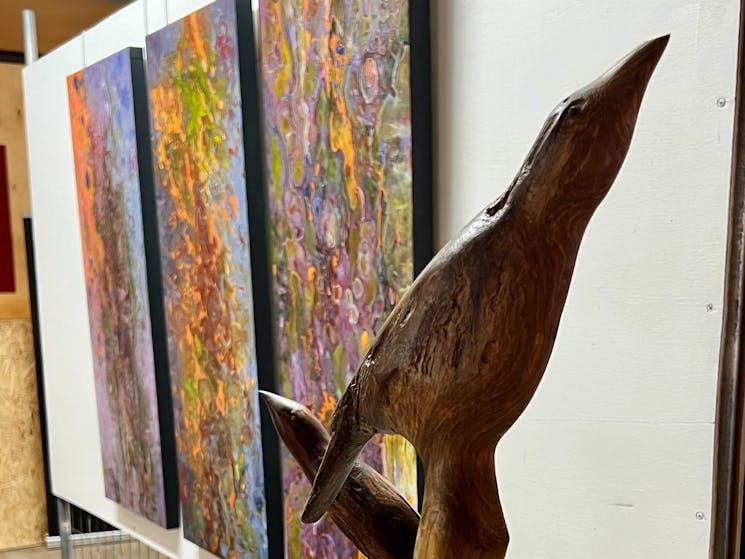 Hand carved wooden birds i on stand in front of a nanging triptych of bark paintings