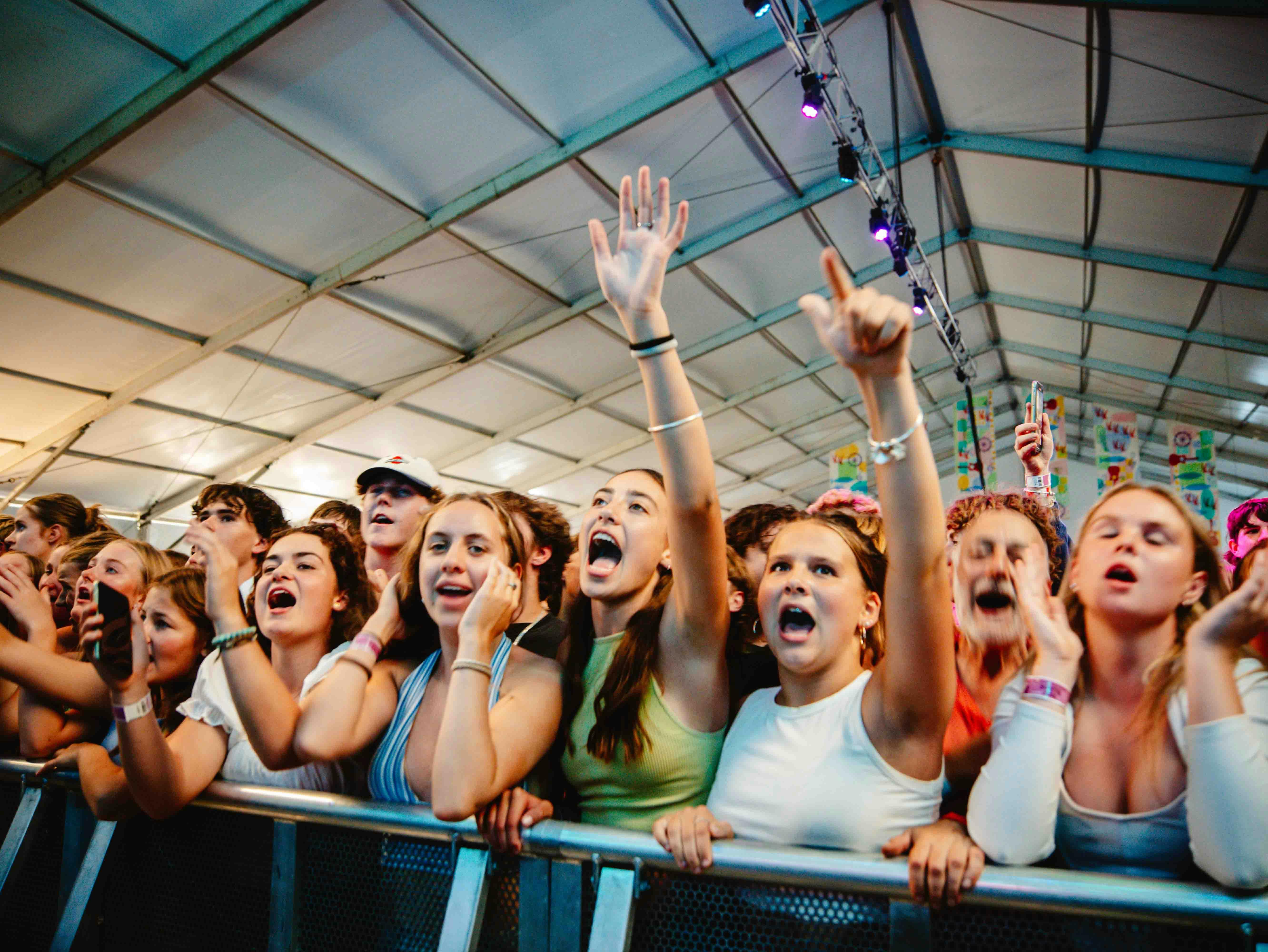 Young people sing along to music against a fence inside a tent. Two people have their arms up.