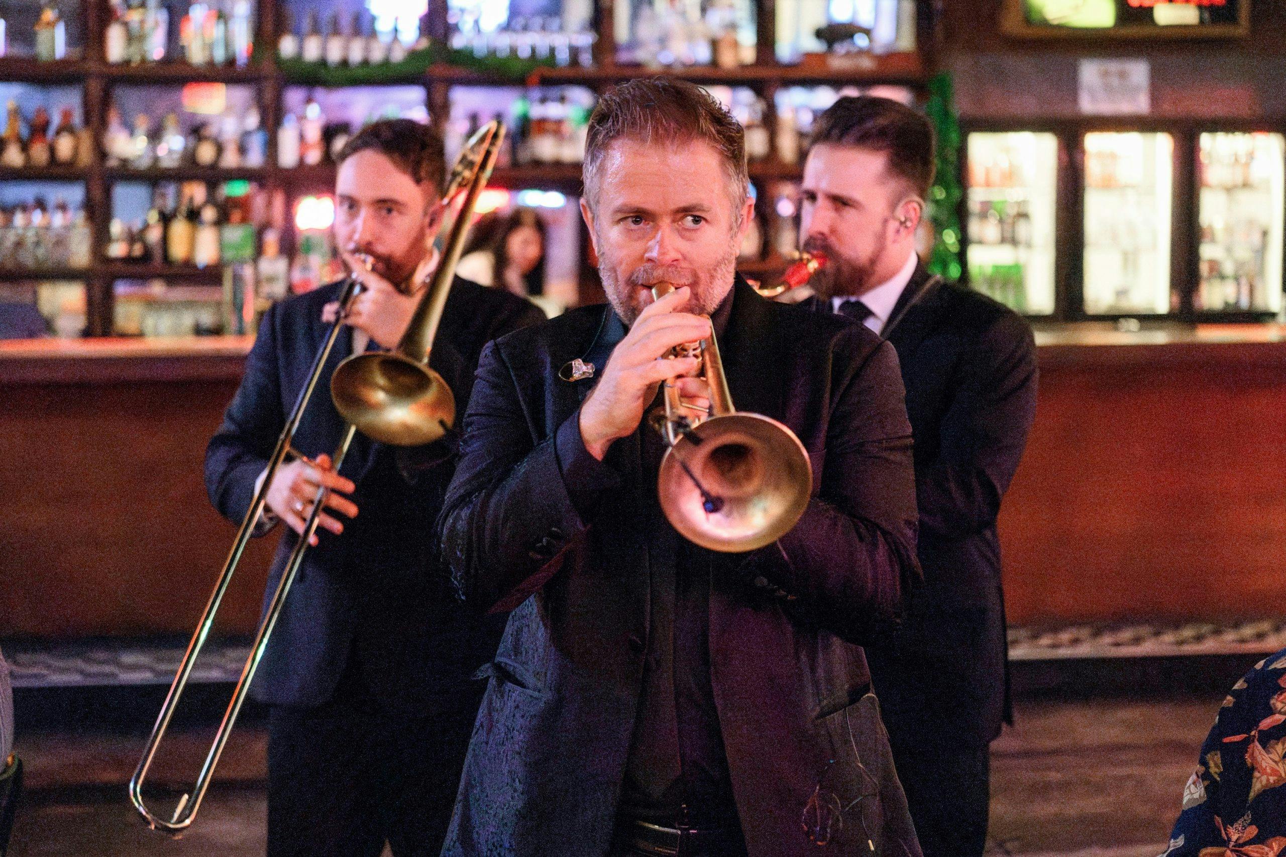 Three suited musicians play brass instruments before a bar.