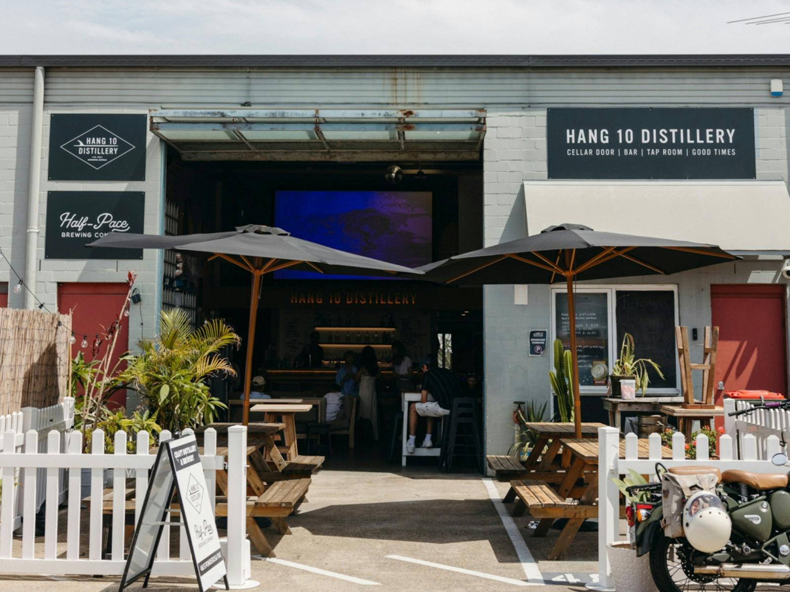 The outside of Hang 10 Distillery with a big door entry, wooden tables, black umbrellas and fences