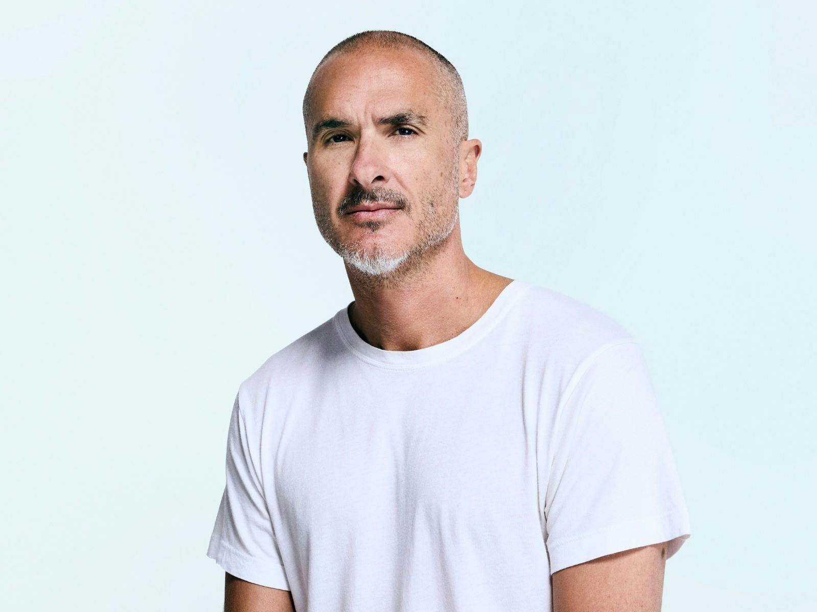 Man in a white t-shirt with a beard staring directly at the camera infront of light blue backdrop.