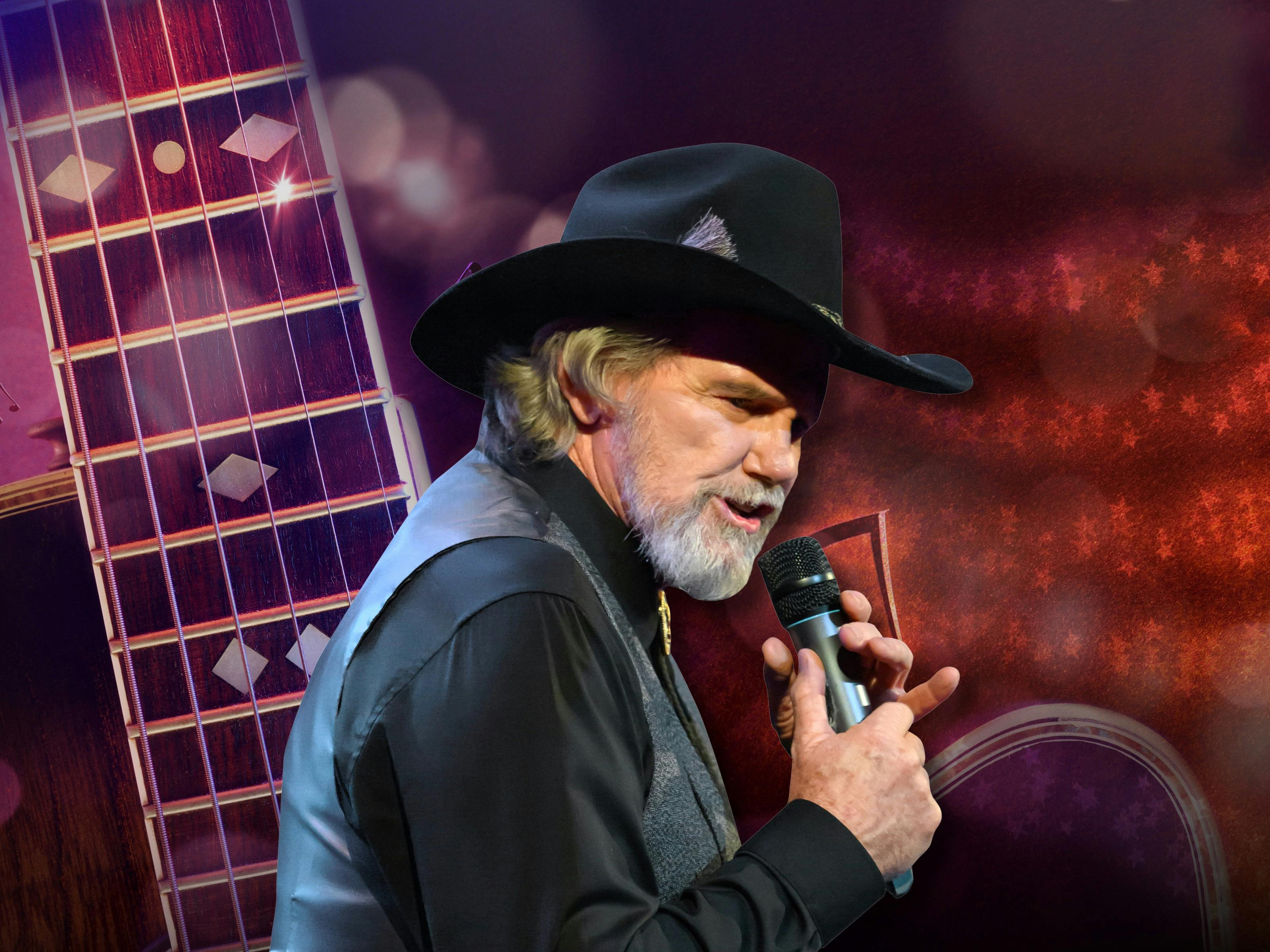 A man in a black outfit and a cowboy hat stands against a coloured background with a microphone