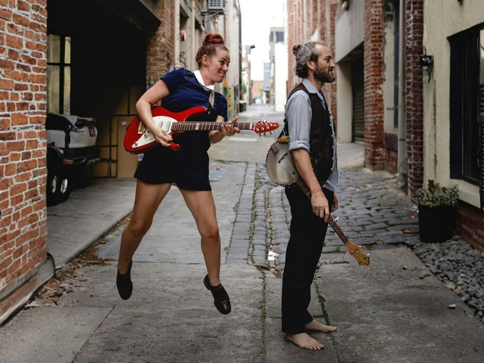 A female jumps holding a red guitar, while a male looks away, they are in a Melbourne laneway.