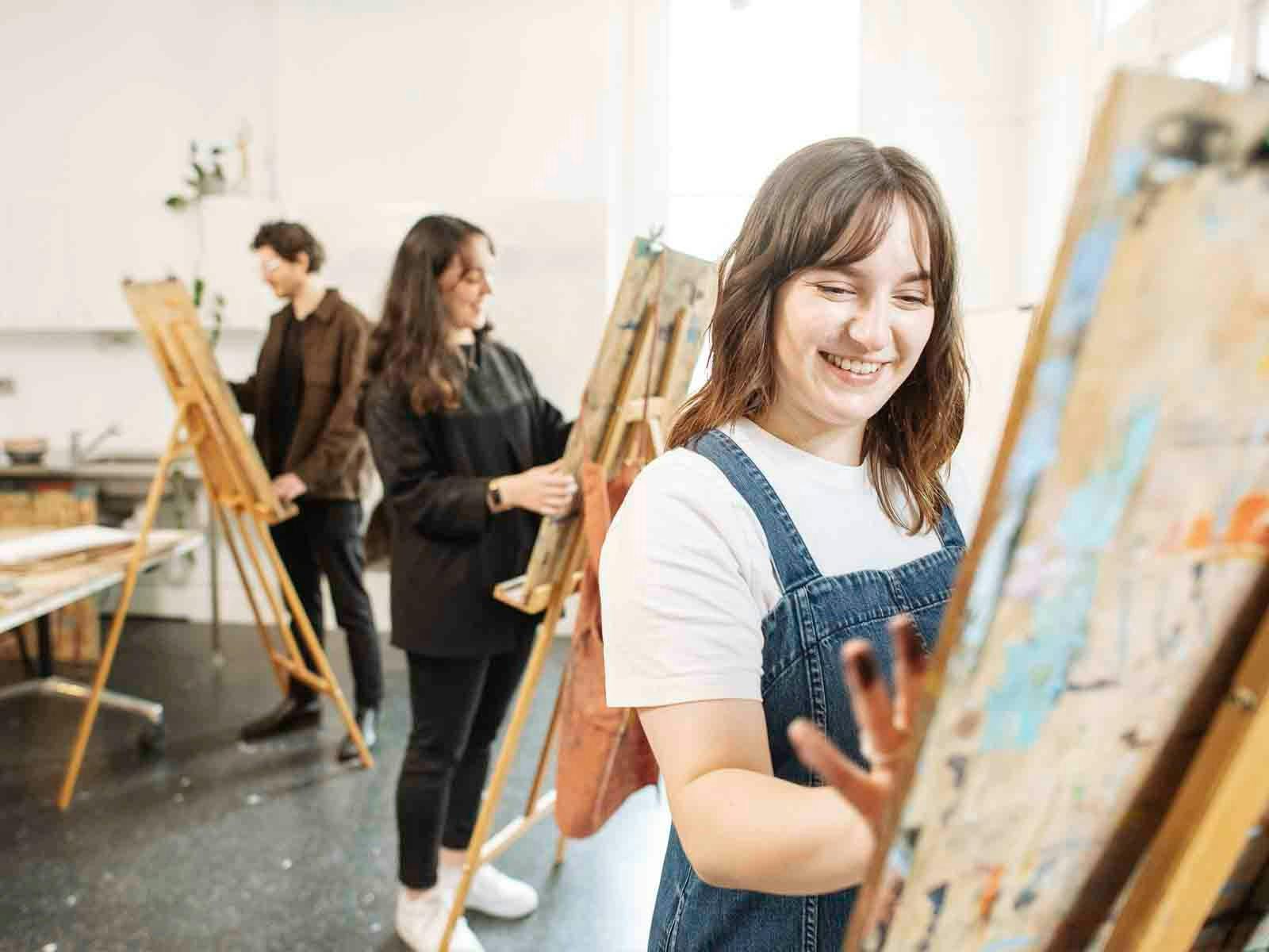 Woman draws at Easle with two other in background of art studio