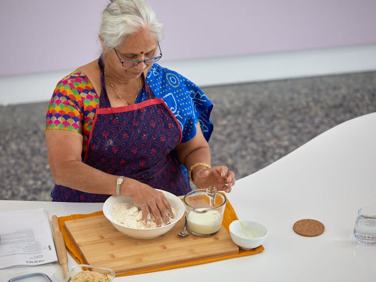 A woman stands at a white kidney bean shaped table mixing flour in a bowl with her hands