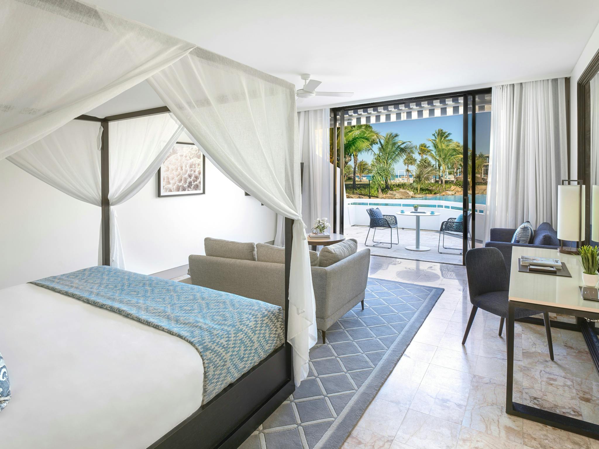 Modern suite featuring a four-poster bed, sitting area, work desk, and balcony overlooking a lagoon Modern suite featuring a four-poster bed, sitting area, work desk, and balcony overlooking a lagoon