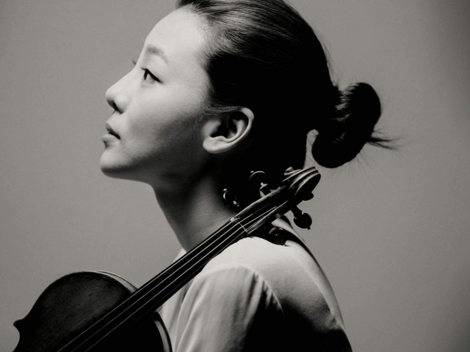 Monochrome photograph of a woman, against a grey backdrop, with a violin leaning on her shoulder.
