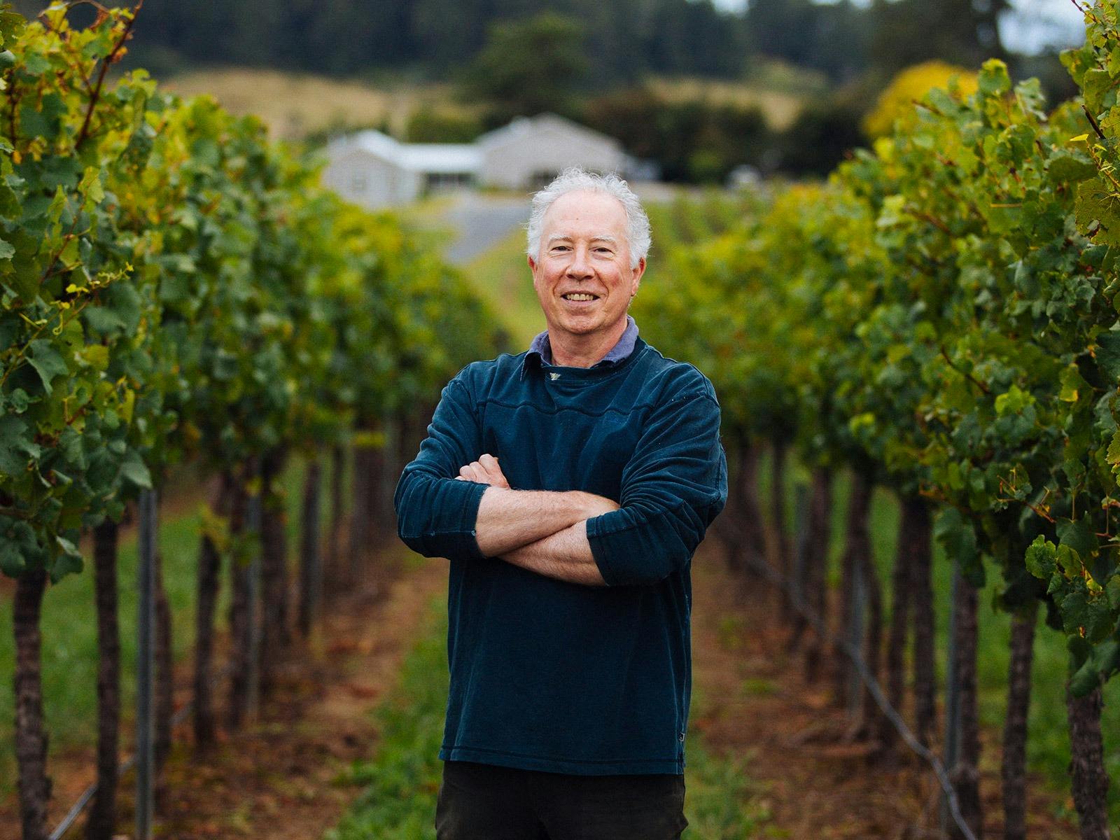 Bill Shrapnel, founder of Colmar Estate, folds his arms and smiles while standing in a row of grapes
