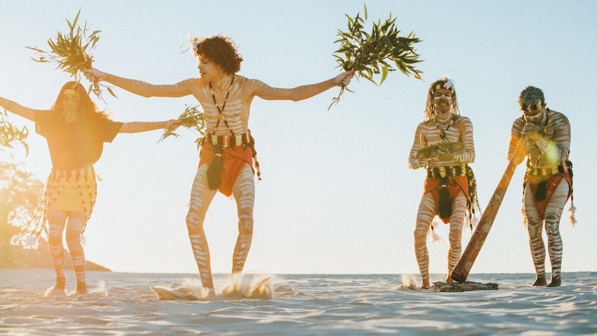 Quandamooka dancers and man playing yidaki on beach, celebrating culture on Minjerribah