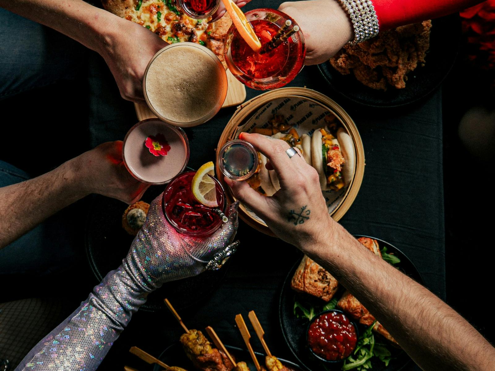 Overhead view of guests raising drinks around a table of bao, pizza and snacks.