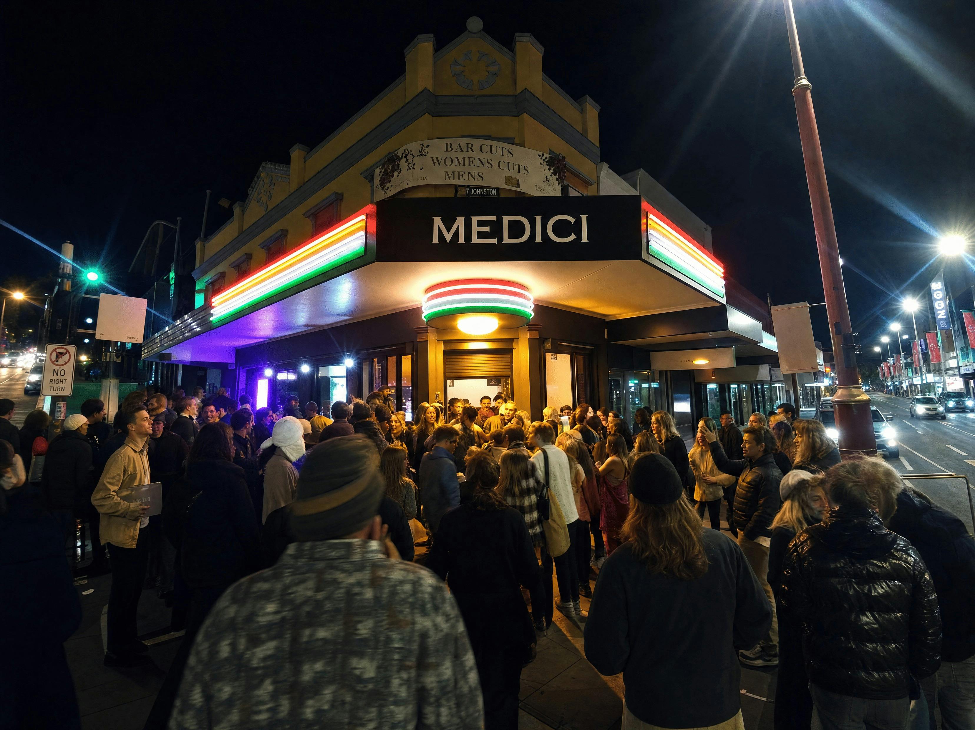 A crowd gathered on the footpath outside the lit              Medici Pizzeria corner entrance at nig