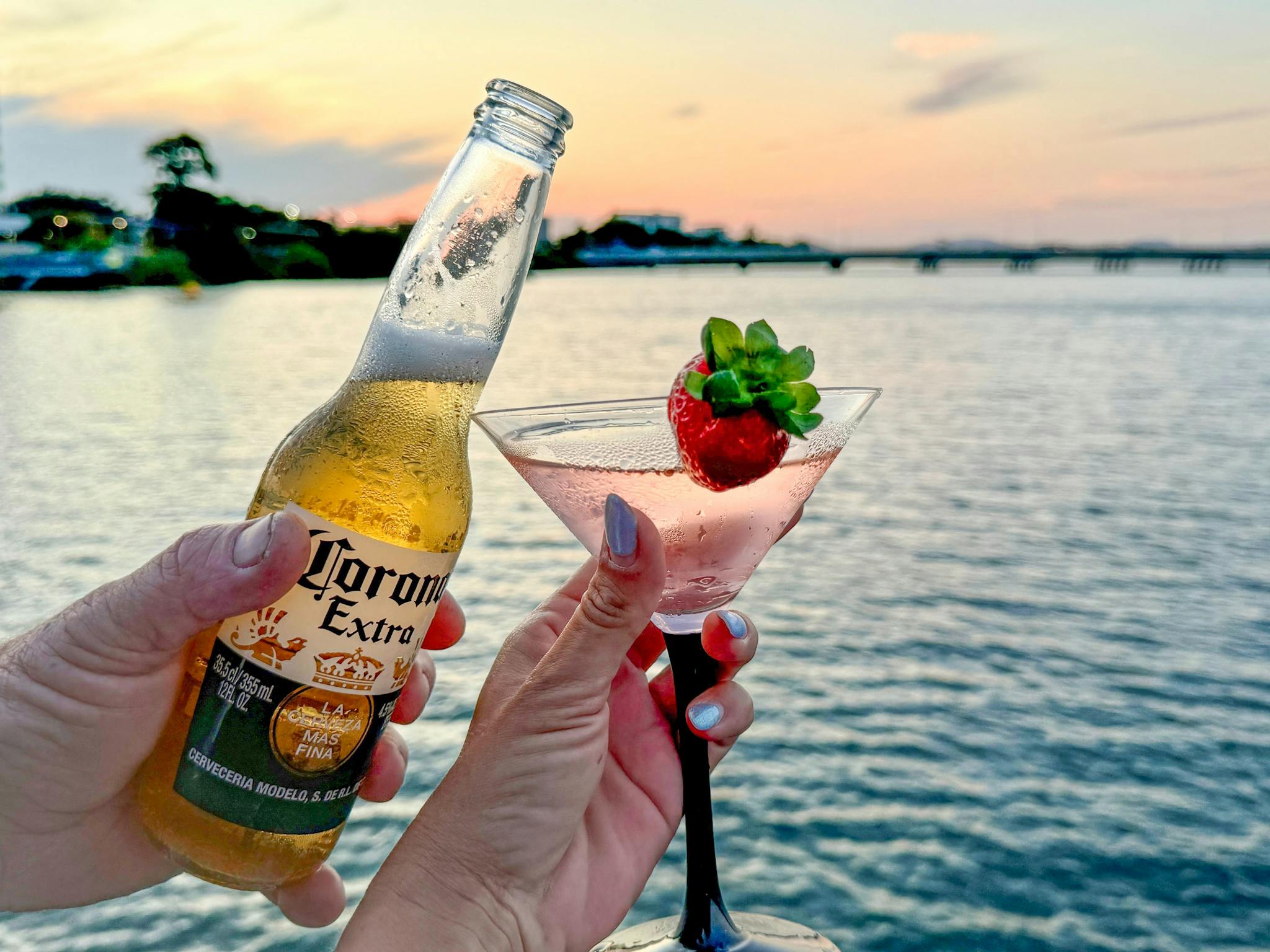 Close-up of two hands holding a beer and a cocktail with a strawberry garnish as the sun sets Close-up of two hands holding a beer and a cocktail with a strawberry garnish as the sun sets