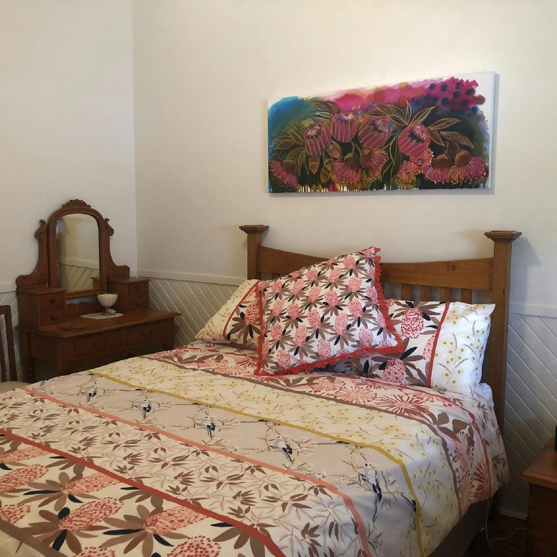 Bed with floral print hanging on wall above bedhead and small dressing table beside bed