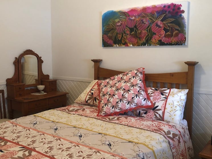 Bed with floral print hanging on wall above bedhead and small dressing table beside bed