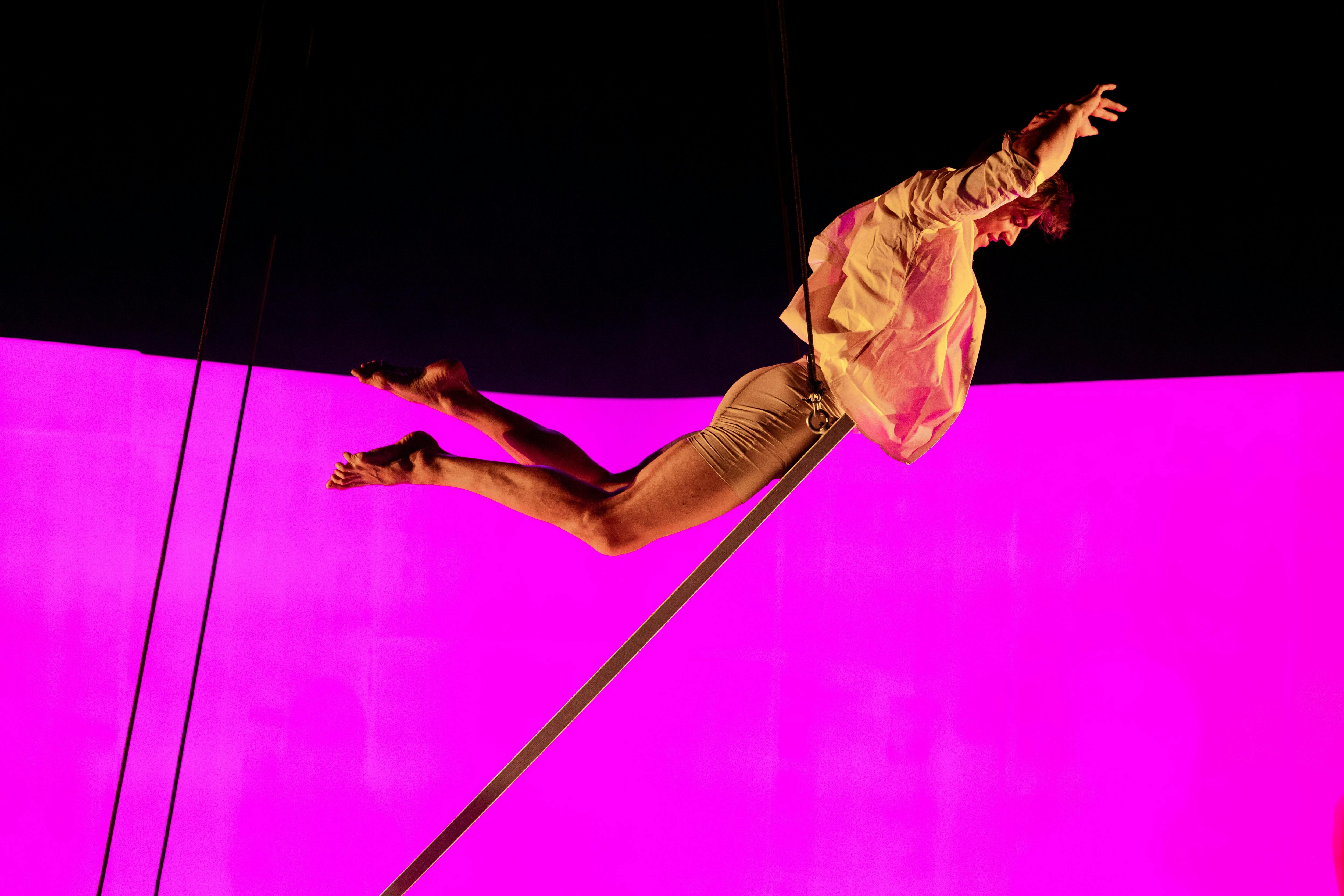A performer wearing an open shirt balances on an inclined board in a joyous moment of flight