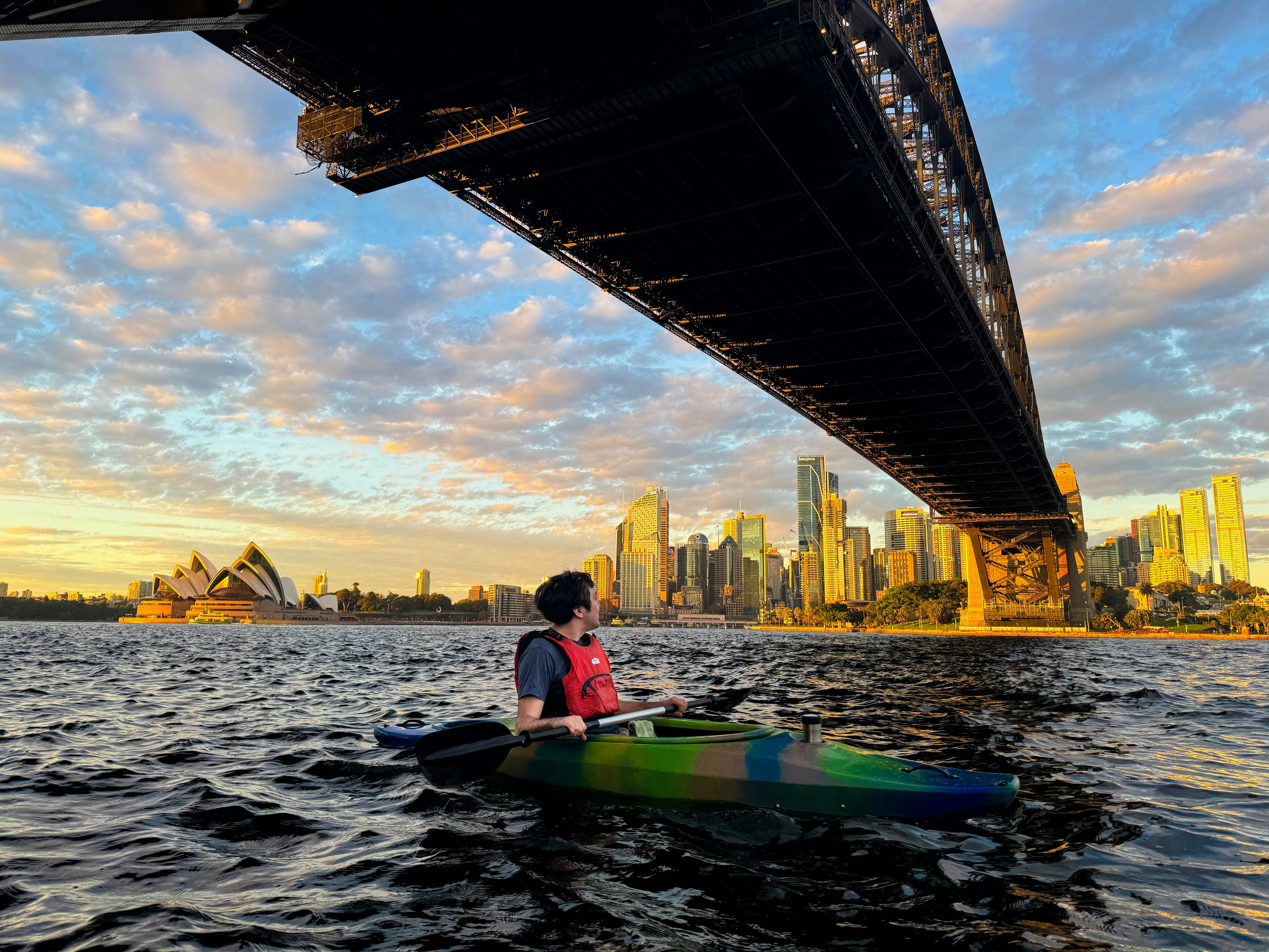 Man kayaks under the Sydney Harbour Bridge with Opera House is background.