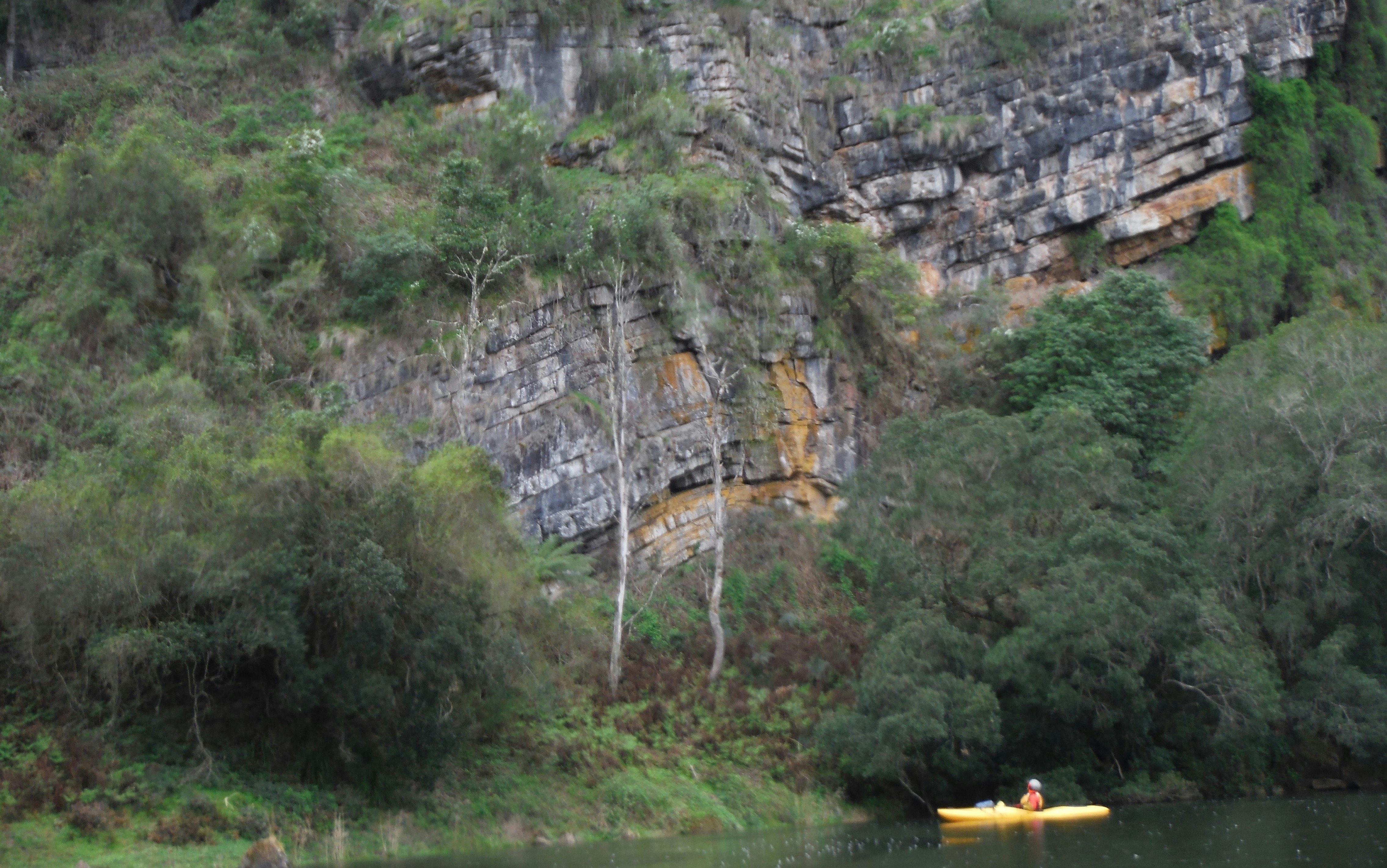 A kayaker is sitting on the snowy river below a wall of ancient different coloured rock layers