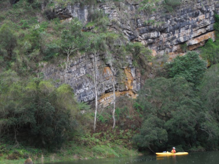 A kayaker is sitting on the snowy river below a wall of ancient different coloured rock layers