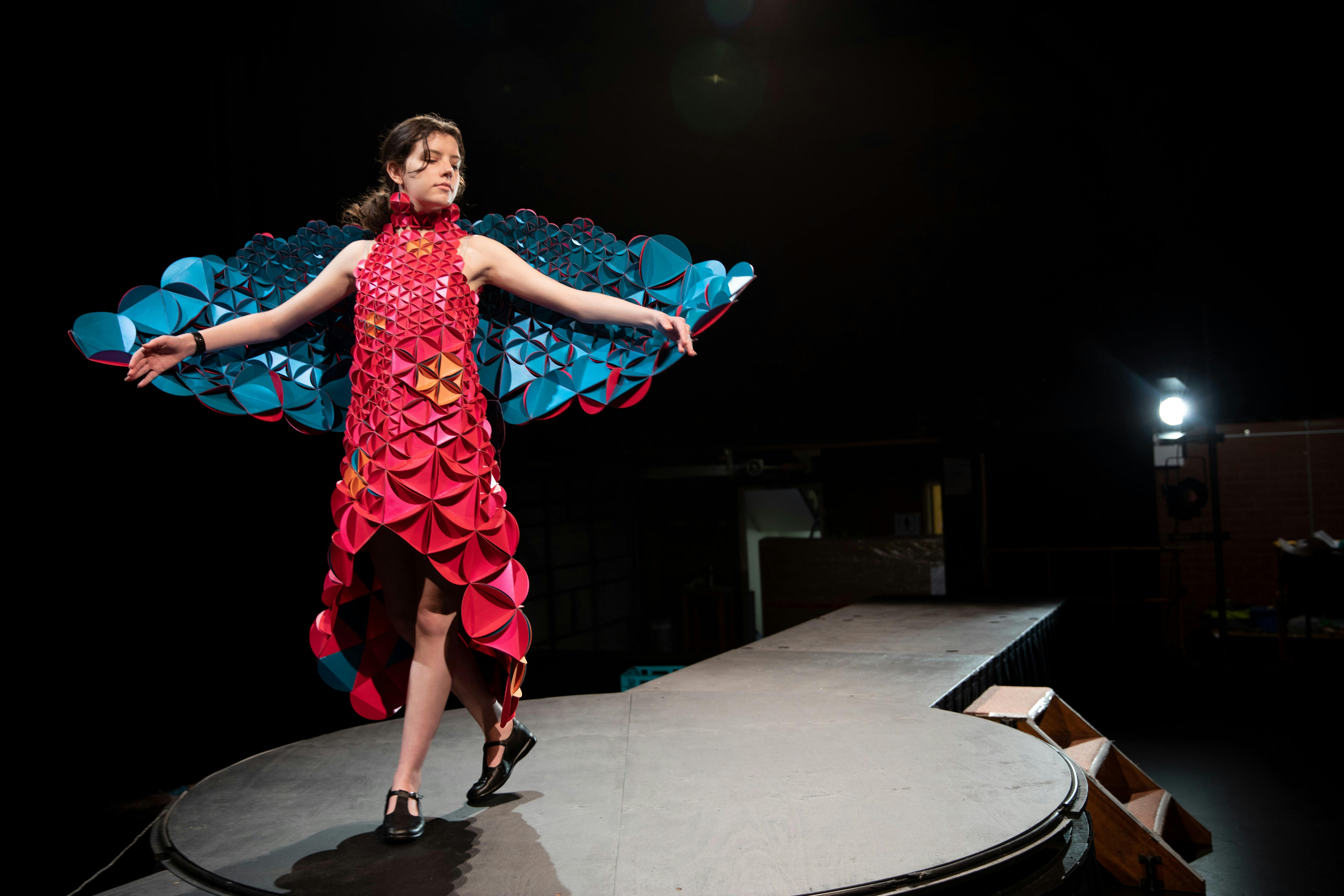 Lady in a pink and teal butterfly dress standing on a stage with her arms outstretched