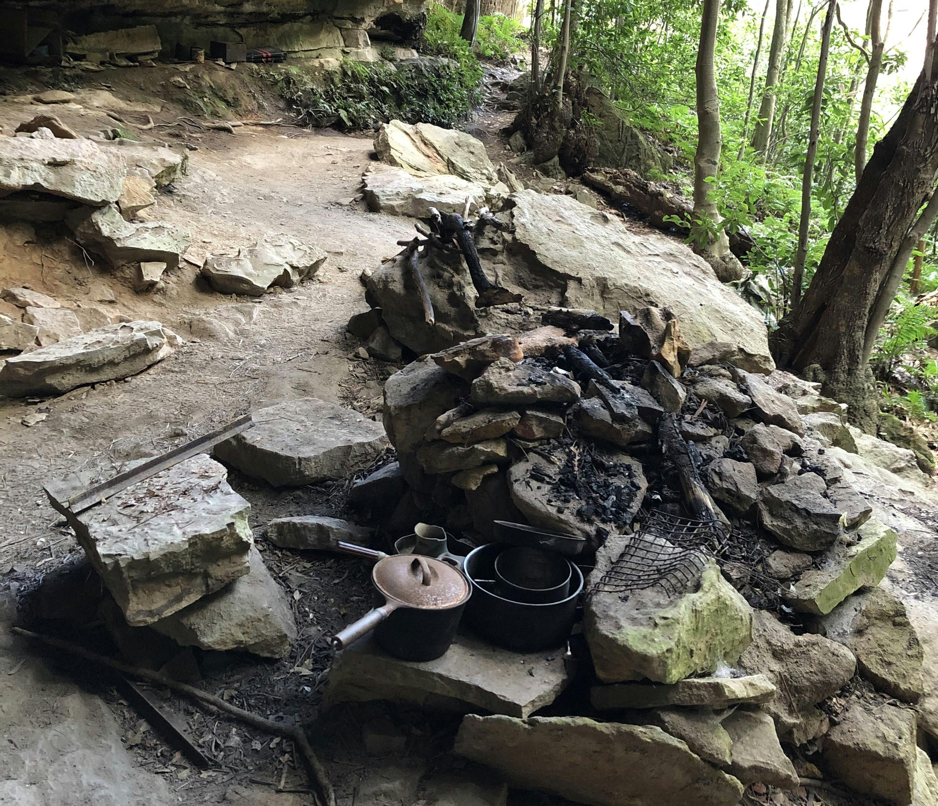 A blackened stone campfire in a sandstone cave, with coachwood trees behine.