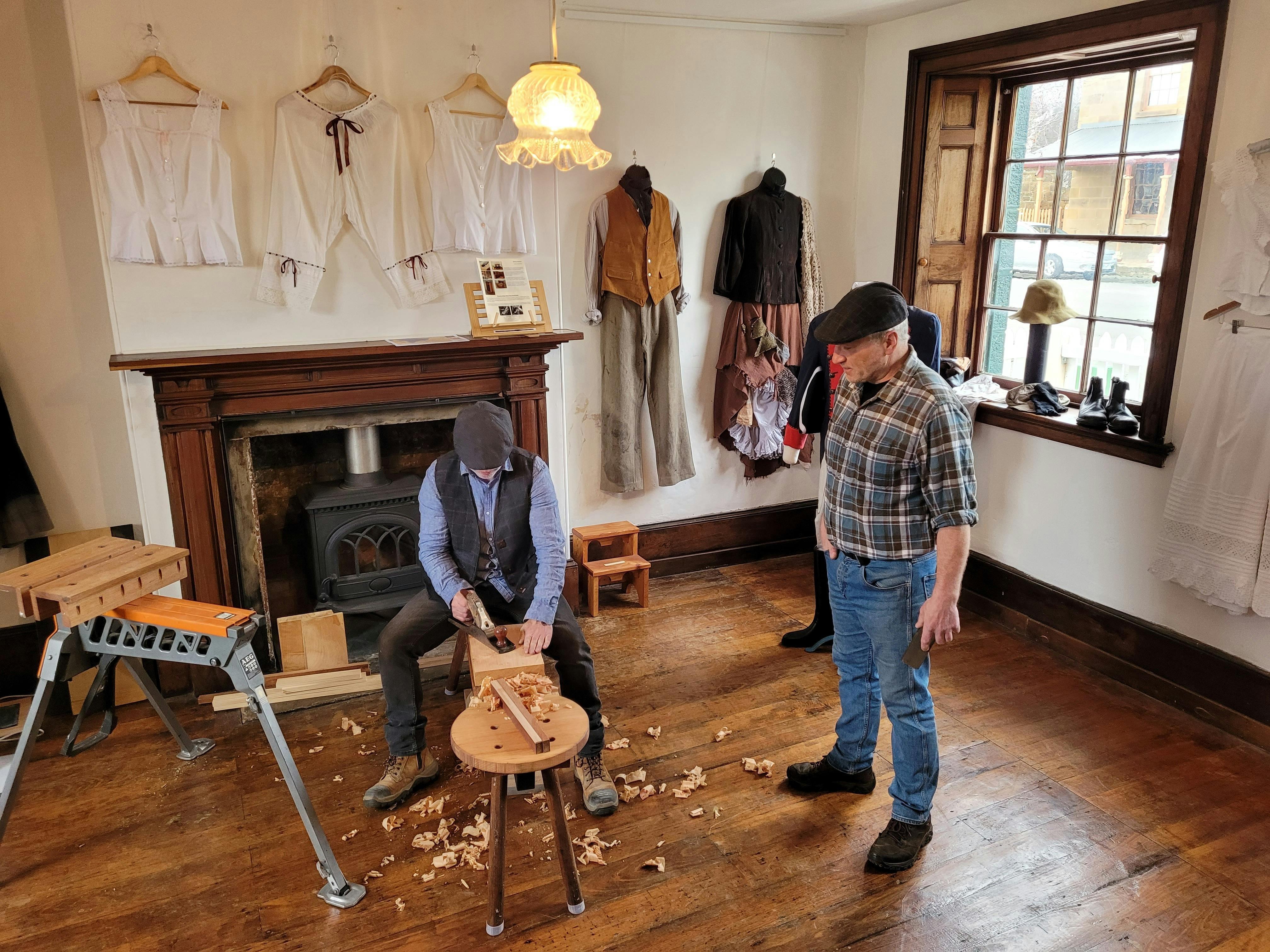 A man uses hand tools to craft a stool leg,another man watches, with historical clothing on the wall