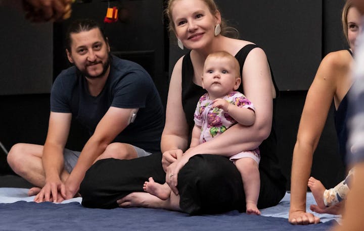 A photograph of 3 adults sitting on the floor, one is holding an infant