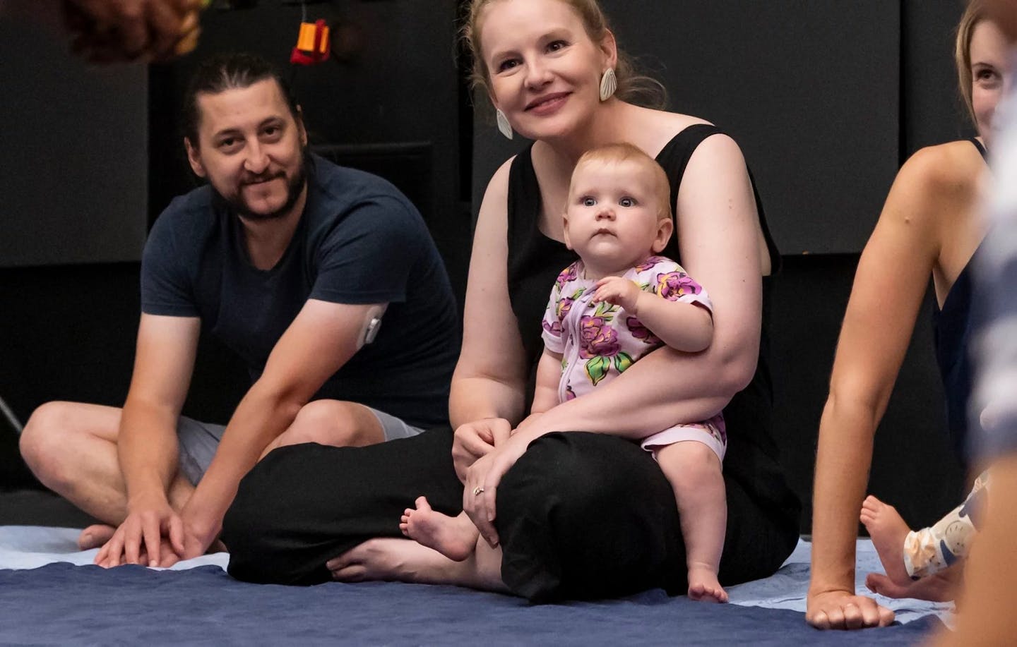 A photograph of 3 adults sitting on the floor, one is holding an infant