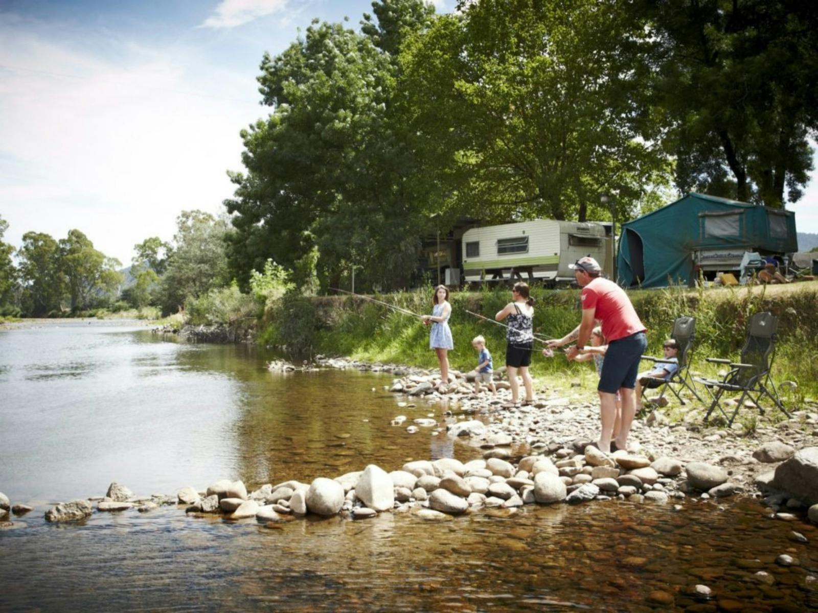 Family of 5 fishing from rocky riverbank, campers and caravans, trees, grass, sunny day
