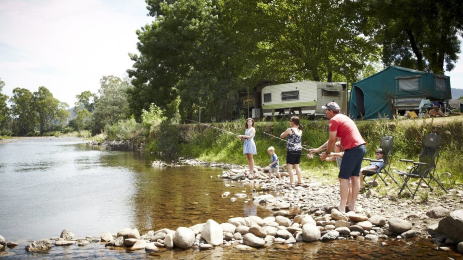Family of 5 fishing from rocky riverbank, campers and caravans, trees, grass, sunny day