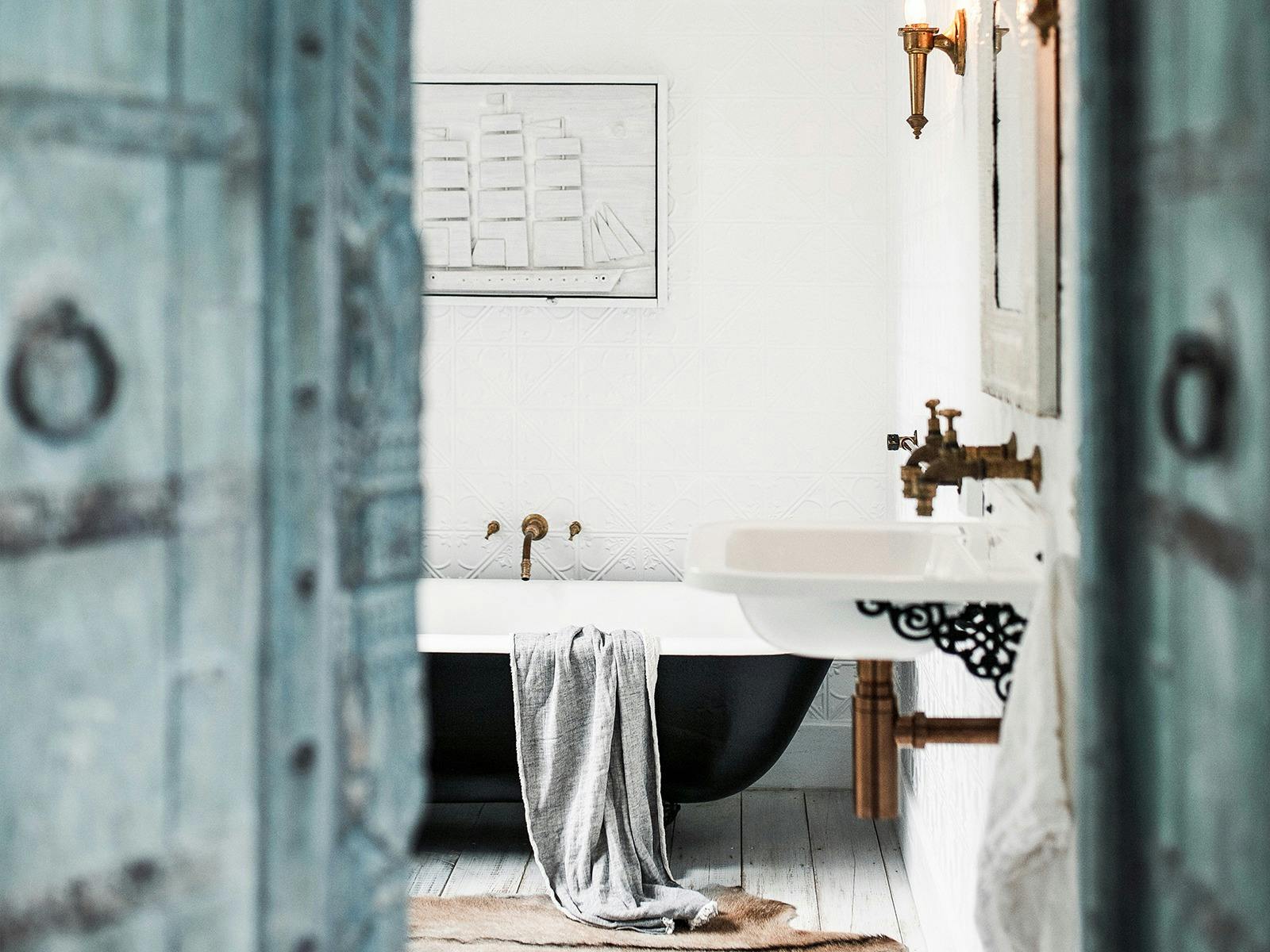 Blue wooden doors lead into the bathroom with a black claw foot bath tub, white wall, gold detailsi
