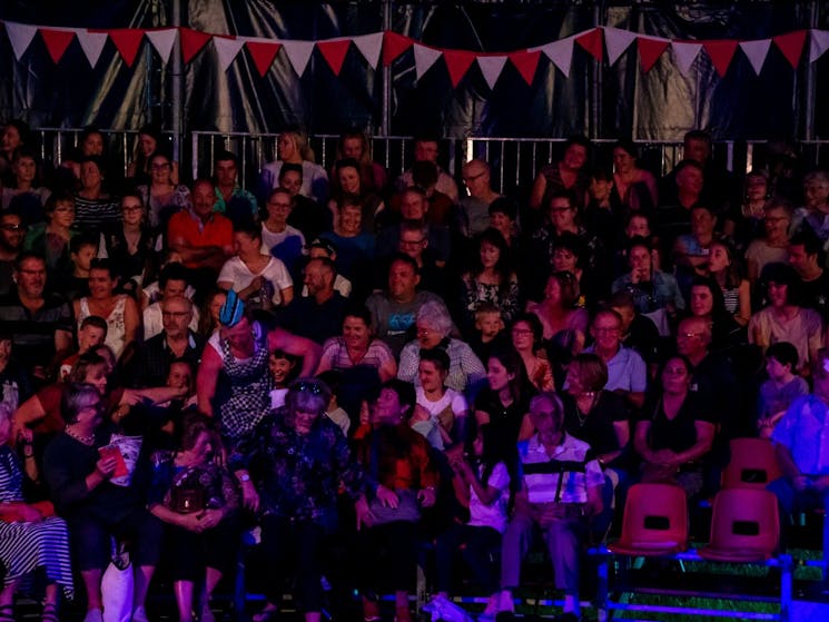 An audience sitting inside a circus big top tent, with colourful bunting hanging overhead.