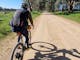Cyclist on right on gravel rd, shadow, green grass, gum trees, blue sky