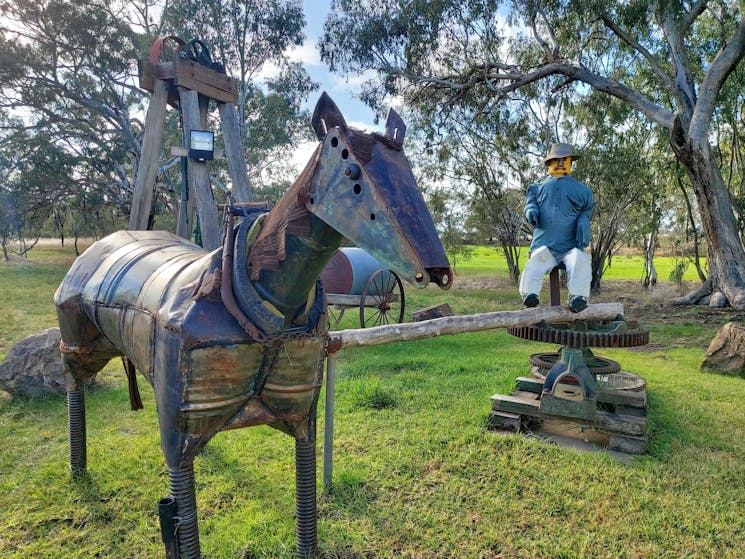 A metal horse sculpture and a man sitting at a water pump in a grassy park.