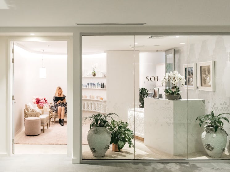 A girl sits in a white reception area reading a treatment menu there is plants and an orchid