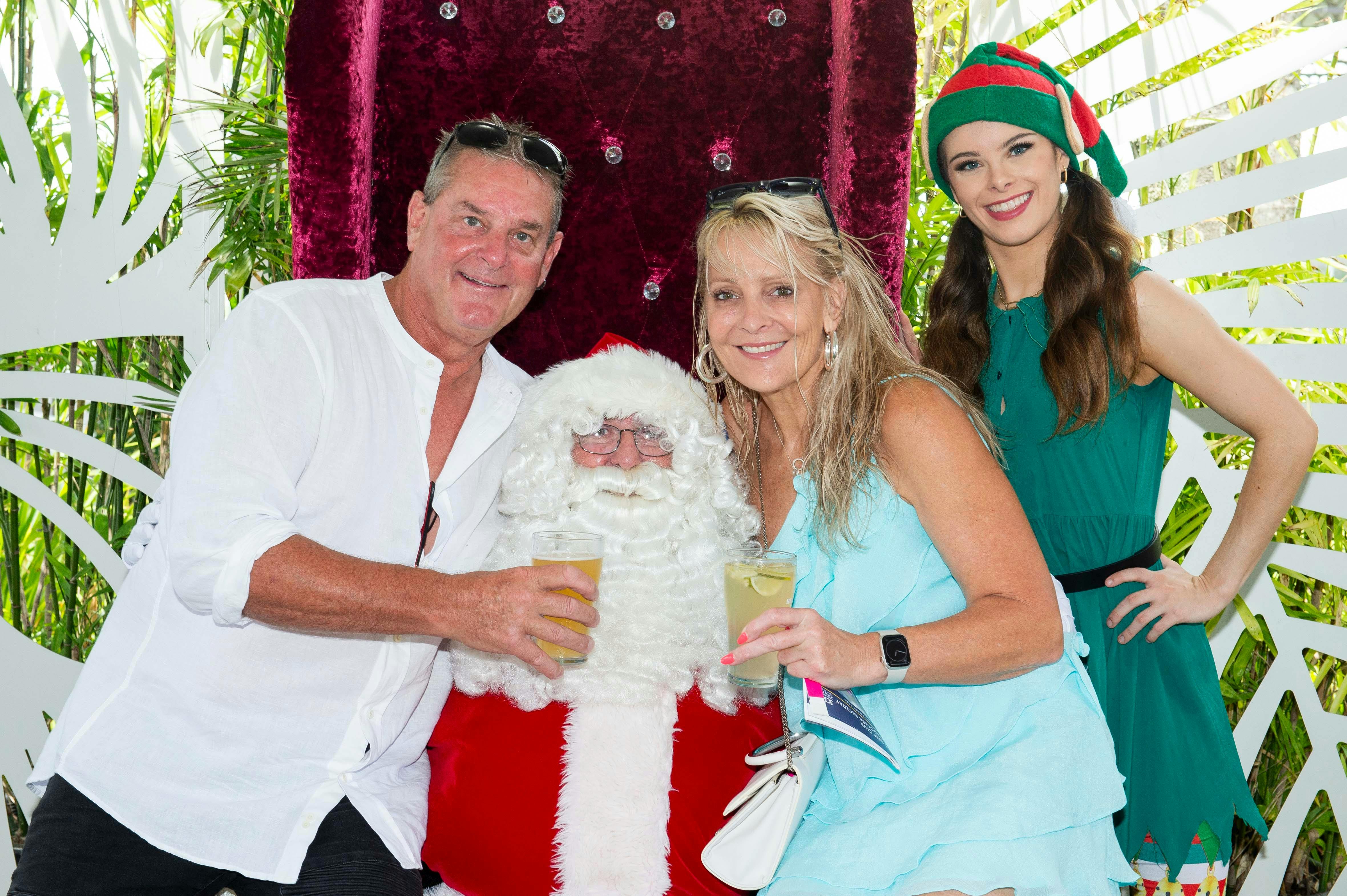 Man and woman holding up beers having their photo with Santa and his elf