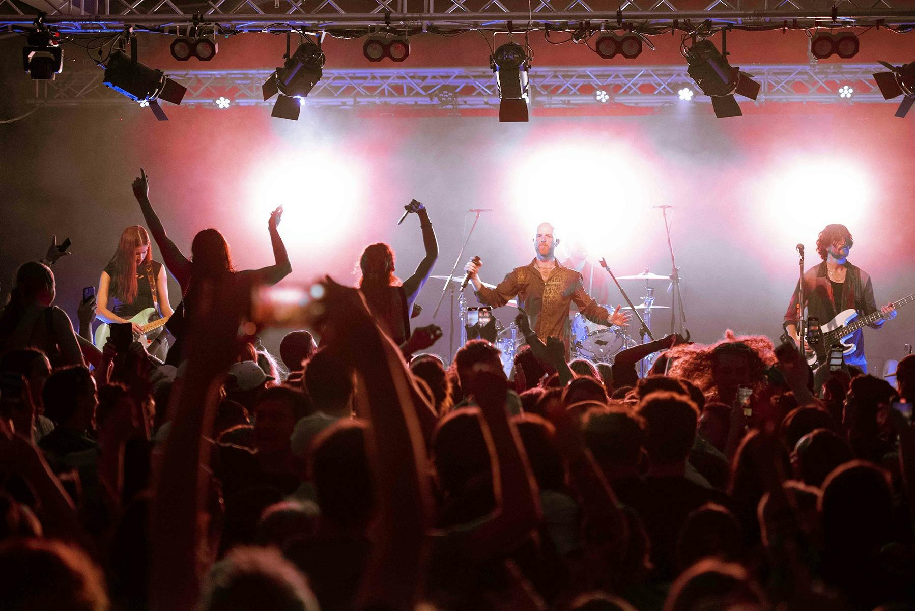 A vibrant rock concert with a lively crowd raising hands. The band on stage, illuminated lights
