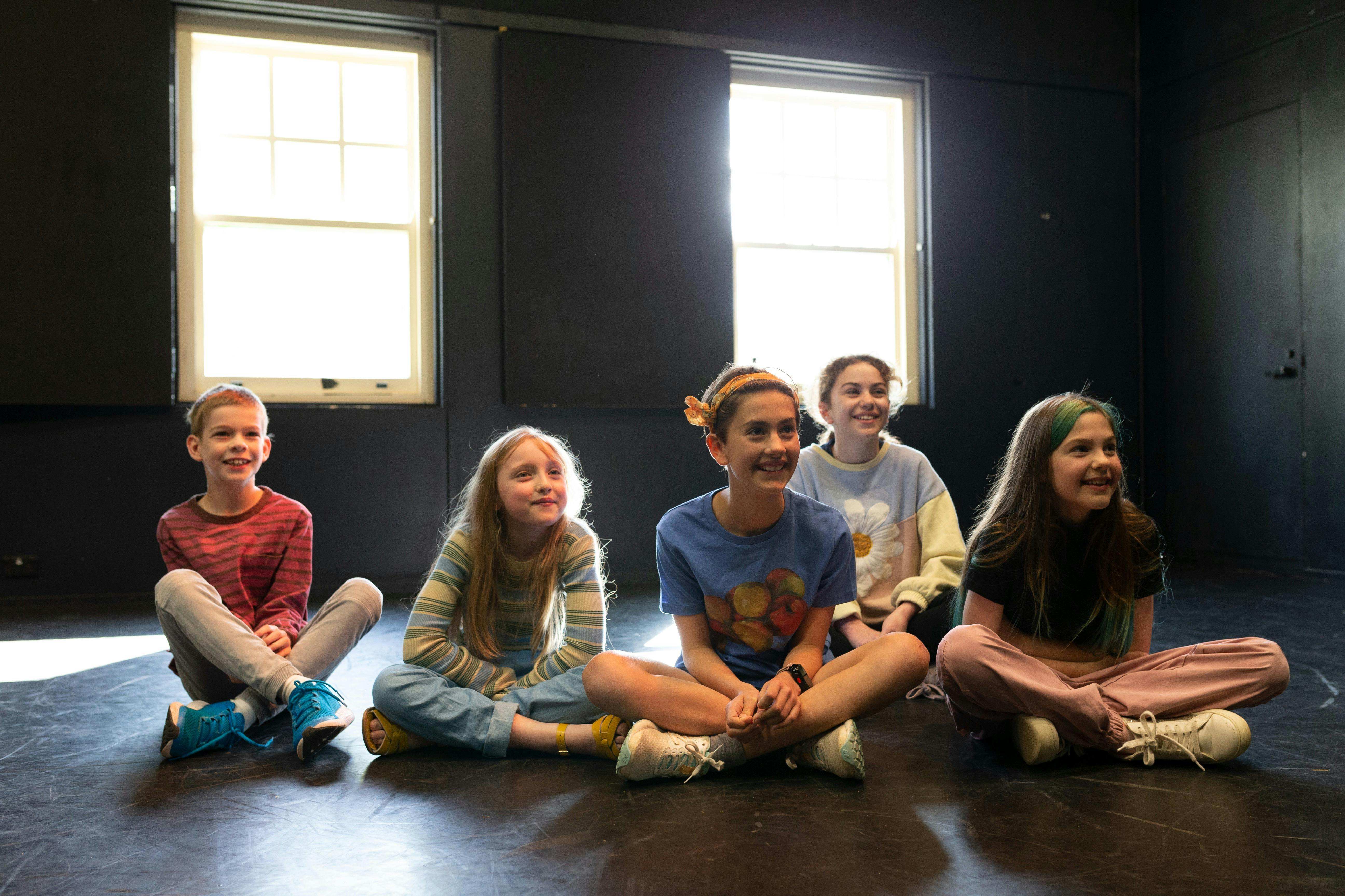 Five children sit cross-legged in a workshop, smiling as they watch a performance.