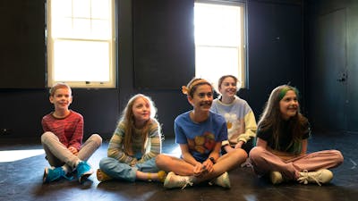 Five children sit cross-legged in a workshop, smiling as they watch a performance.
