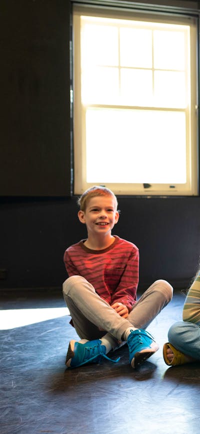 Five children sit cross-legged in a workshop, smiling as they watch a performance.