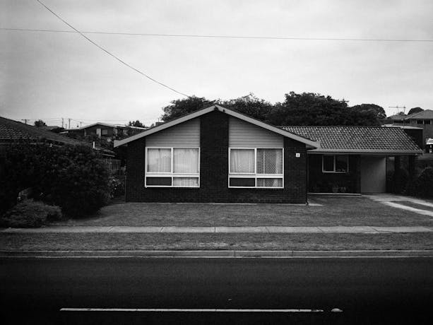 A black and white photograph of a suburban scene featuring a house, a bare front yard and street