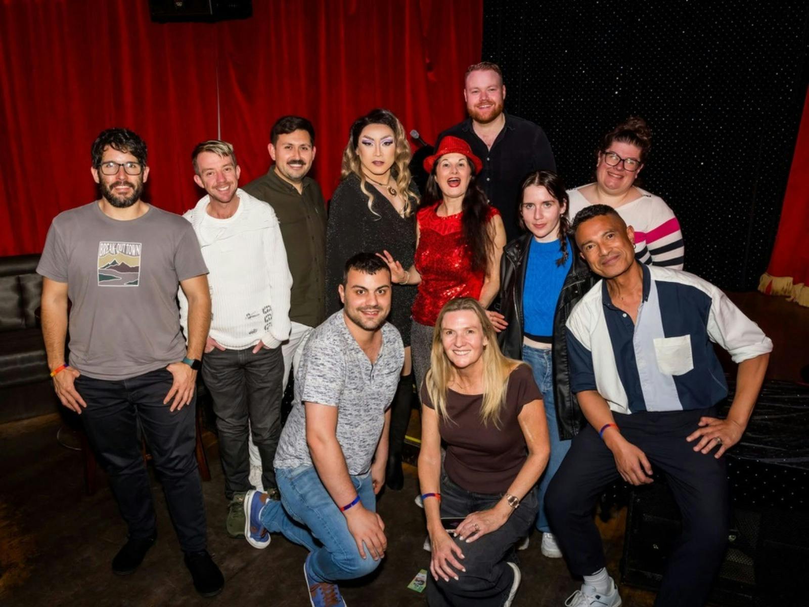 A group of performers posing together in front of red stage curtains.