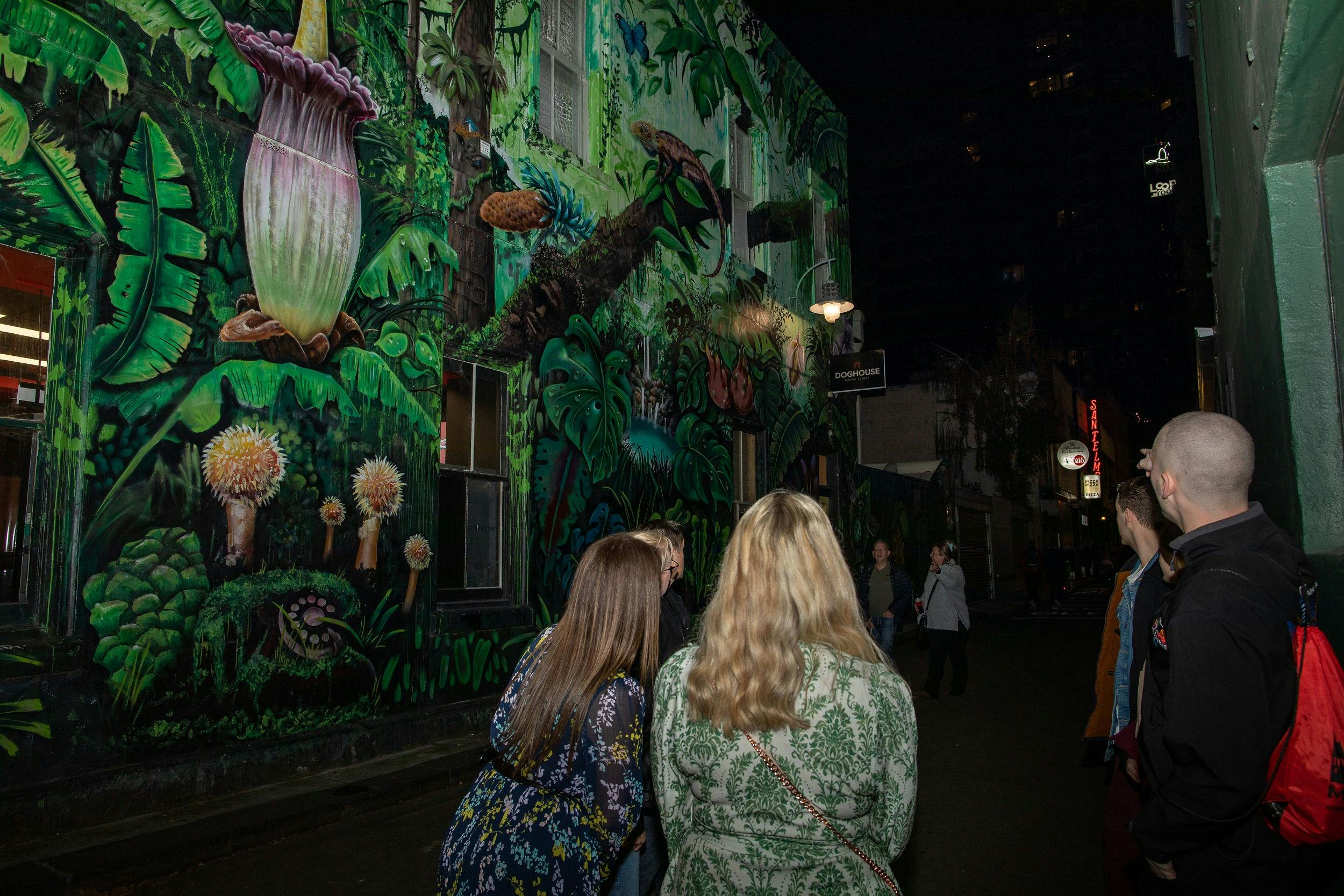 A small group of people standing in a darkened lane.  There is a large botanical mural on one wall.